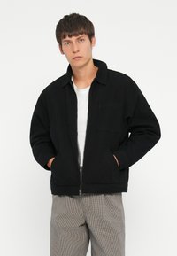 Black zip-up jacket with a collar, two side pockets, and a single chest pocket. Worn over a white t-shirt and paired with plaid pants.