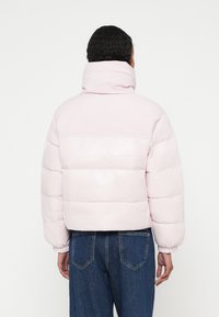 Persoană care poartă o geacă matlasată roz deschis cu guler înalt și manșete elastice, asortată cu blugi din denim albastru închis, văzută din spate.