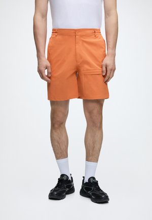 Muž oblečený v oranžových cargo šortkách, bielych ponožkách a čiernych teniskách stojaci na jednoduchom bielom pozadí.