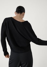 Personne aux cheveux attachés en chignon portant un pull noir ample tombant d'une épaule et un pantalon noir, vue de dos sur un fond uni.