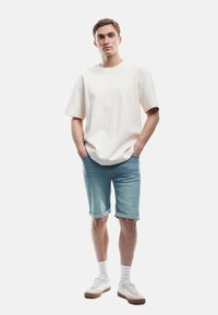 Joven de pie con las manos en los bolsillos, vistiendo una camiseta blanca de gran tamaño, pantalones cortos de mezclilla azul claro enrollados, calcetines blancos y zapatillas blancas.