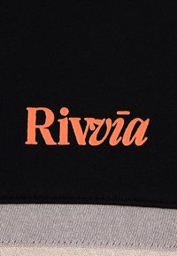 Μαύρο ύφασμα με κεντημένο πορτοκαλί λογότυπο «Rivviā». Κοντινή προβολή που δείχνει λεπτομέρειες υφής και ραφής, σε αντίθεση με το λείο φινίρισμα του υφάσματος.