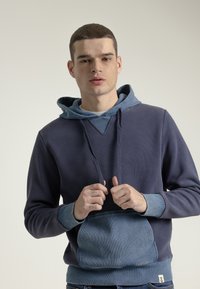 Sudadera azul marino con una parte superior texturizada y un bolsillo canguro de un azul más claro. Presenta una capucha en contraste y puños acanalados. Diseño simple.