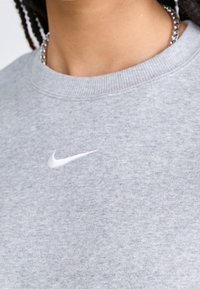 Harmaa huppari, jossa on urheilullinen pyöreä kaula-aukko ja pieni valkoinen Nike-logo rinnassa. Materiaali näyttää pehmeältä ja teksturoidulta.