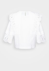 Blouse blanche avec des manches en dentelle à œillets en forme de coquille et un ourlet en dentelle. Design court et ajusté avec une ouverture en forme de clé dans le dos pour le détail de la fermeture.