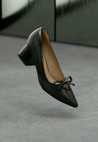 Μαύρο δερμάτινο γυναικείο παπούτσι με χαμηλό τακούνι, μυτερή μύτη, γυαλιστερή διακόσμηση και λεπτομέρεια φιόγκου, τοποθετημένο πάνω σε γκρι μοκέτα.