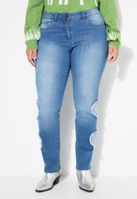 Pantalones vaqueros de mezclilla azul desgastados con cortes desgastados en las rodillas, ajuste slim y diseño de cinco bolsillos. Combinados con botas de tobillo metálicas plateadas.