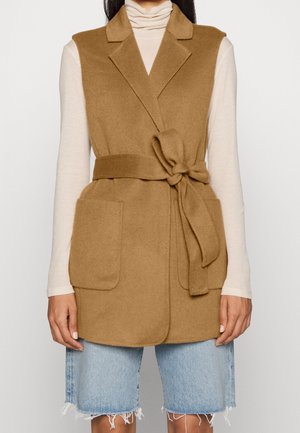 Bezrękawnik z wełny w kolorze wielbłądzim brązie z paskiem, noszony na kremowym golfie, zestawiony z jasnoniebieskimi, dżinsowymi, postrzępionymi szortami do kolan.