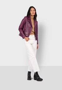 Žena v purpurové kožené bundičce, béžovém roláku, bílých kalhotách a černých botách na podpatku, stojící na světle šedém pozadí.