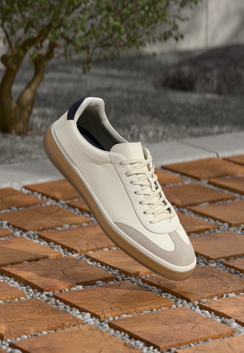 Witte leren sneaker met grijze suède accenten, ronde neus en beige rubberen zool, met een klassieke vetersluiting en marineblauwe hiel detail.