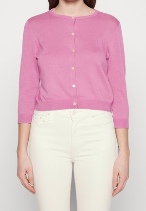 Mujer que lleva un cárdigan de punto rosa con botones, mangas tres cuartos y pantalones de cintura alta en color blanco roto.