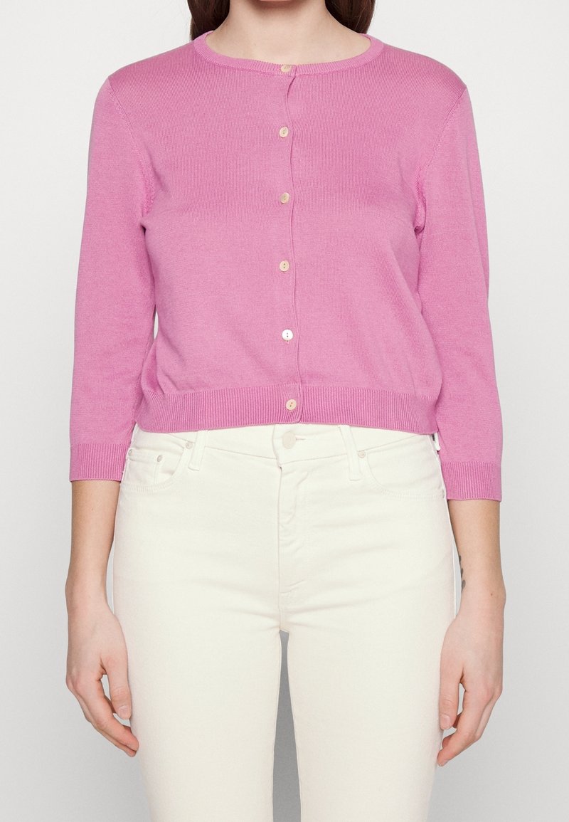 Femme portant un cardigan en tricot rose boutonné avec des manches trois-quarts et un pantalon taille haute couleur écru.