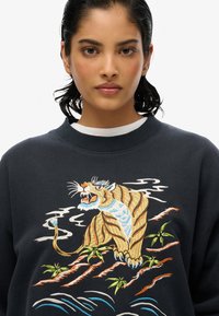 Sudadera gris oscuro con un colorido diseño de tigre bordado, acompañado de bambú y olas. Tiene cuello redondo, mangas largas y tejido texturizado.