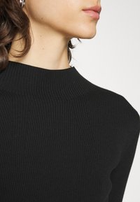 Pull en maille côtelée noir avec un col montant et de subtils détails texturés, mettant en avant une coupe ajustée et un tissu doux.