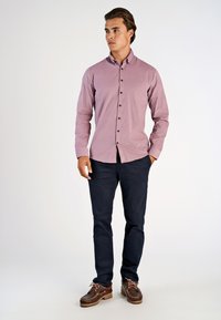 Camicia button-up rosa chiaro con colletto e bottoni neri, abbinata a pantaloni blu scuro e scarpe marroni con lacci. Abbigliamento casual in mostra.