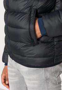 Tengerészeti puff jacket vízszintes varrással, cipzáras zsebekkel és kontrasztos kék díszítéssel a mandzsettákon. Anyaga simának és szigeteltnek tűnik.