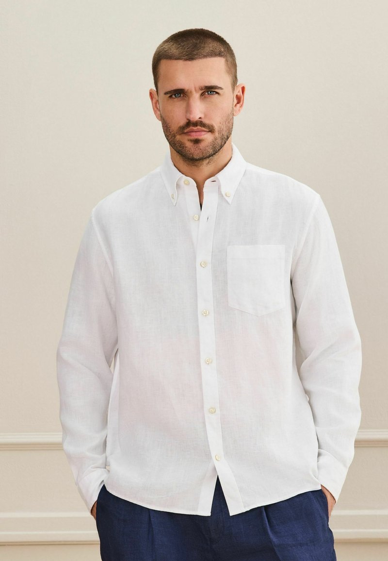 Homme aux cheveux courts et à la barbe portant une chemise blanche boutonnée et un pantalon bleu foncé, debout contre un mur beige.
