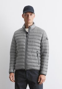 Grå oppustelig jakke med horisontal quiltet design, høj krave og lynlåslommer. Kombineret med en mørk kasket og en simpel hvid t-shirt med runde halsudskæring.
