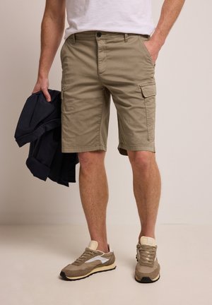 Mies, jolla on beigeiset cargo-shortsit ja vaaleanruskeat tennarit, pitelee vasemmassa kädessään tummaa takkia, oikea käsi on taskussa neutraalia taustaa vasten.