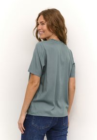 Trumpos rankovės blusa prislopinto smaragdo spalvos su minkšta tekstūra, su raukšlėta apykakle. Sujungta su tamsiai mėlynos džinsinės kelnių.