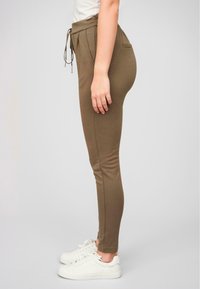 Smalte olivenfargede joggebukser laget av myk stoff med elastisk linning, snøring og sidelommer, kombinert med hvite sneakers.