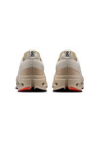 Beige athletic shoes with a textured upper, prominent cushioning, and a black and orange sole. Features a logo on the heel and sleek design.