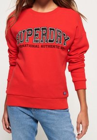 Sudadera roja con cuello redondo y puños acanalados. Presenta un gran texto negro "SUPERDRY" y una pequeña etiqueta de marca en el dobladillo.
