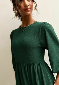 Grünes, geripptes Kleid mit rundem Ausschnitt, gepufften Ärmeln und ausgestelltem Rock. Es zeichnet sich durch ein strukturiertes Material und eine glatte Oberfläche aus.