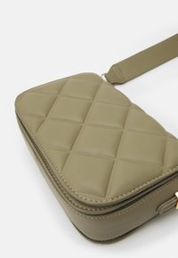 Olivově zelená crossbody taška s prošívaným polstrovaným vzorem, zakulacenými okraji a nastavitelným popruhem na bílém pozadí.