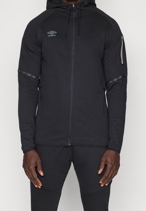 Uomo che indossa una felpa nera con cappuccio e zip, logo Umbro, tasca con zip sulla manica sinistra e pantaloni neri abbinati.