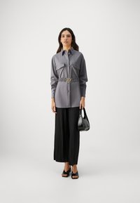 Chemise grise à boutons avec deux poches poitrine et une taille ceinturée, associée à un pantalon large plissé noir et des chaussures ouvertes noires.