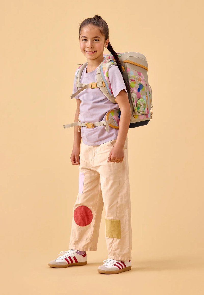 Rugzak met een kleurrijk stippenpatroon, beige banden; lichtpaarse T-shirt; crèmekleurige broek met rode, roze, gele vlekken; witte sneakers.