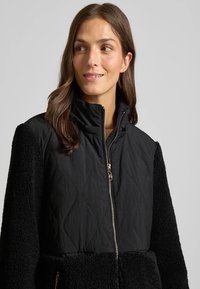 Zwarte zip-up jasje met een gewatteerd bovenstuk en tekstuursweater mouwen. Bevat een hoge kraag en gouden hardwareaccenten.