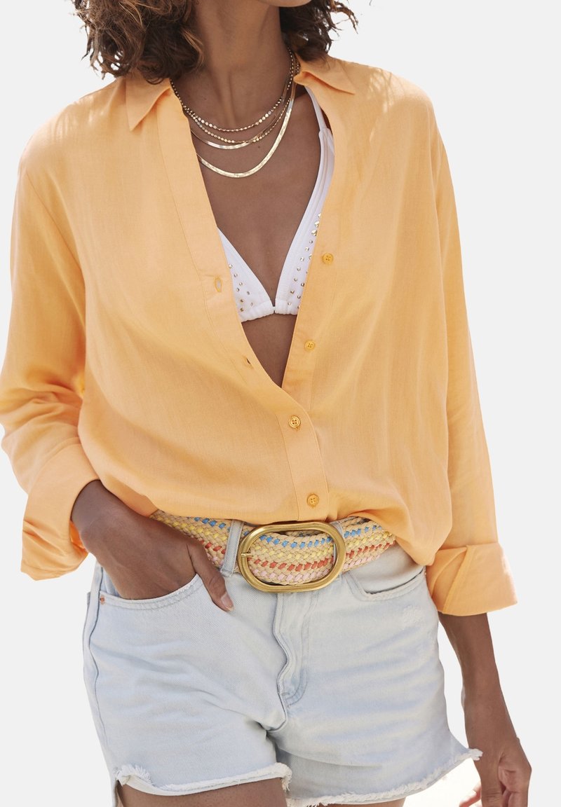 Femme portant une chemise jaune ouverte à boutons sur un haut de bikini blanc clouté, un short en jean clair, une ceinture tissée multicolore et des colliers dorés superposés.