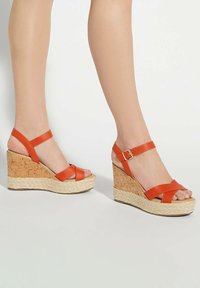 Orange leather wedge sandals with crossed straps and ankle buckle, featuring a textured cork and rope platform.