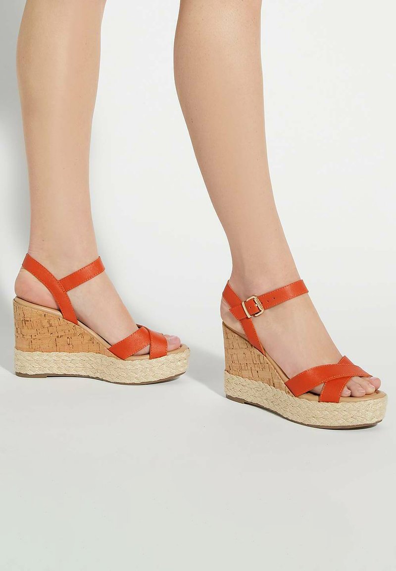 Orange leather wedge sandals with crossed straps and ankle buckle, featuring a textured cork and rope platform.