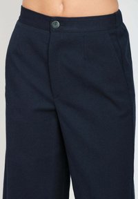 Pantaloni blu navy con una vita con bottoni e tasche laterali, indossati da una persona con la pancia nuda visibile.