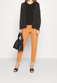 Cárdigan ligero negro sobre una camiseta negra, combinado con pantalones naranja ajustados. Un bolso negro fruncido y sandalias negras de dedo completan el look.