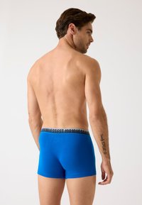 Zilie bokseršorti ar melnu logo jostas daļu, kas piedāvā ciešu piegulšanu un gludu tekstūru. Modelis attēlots no aizmugures, bez augšdaļas.