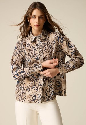 Vrouw met lang bruin haar, gekleed in een beige overhemd met donkerblauwe abstracte patronen en een witte broek, terwijl ze haar mouw rechtzet.