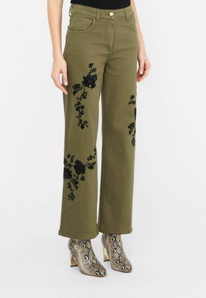 Olivegrüne, weit geschnittene Hose mit schwarzer Blumenstickerei, kombiniert mit schlangenledermuster-Absatz-Stiefeletten und einem schwarzen Oberteil.