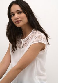 Blusa bianca con un corpetto in pizzo, maniche a cappuccio e un dettaglio a bottoni sul colletto. Tessuto morbido con una texture liscia.