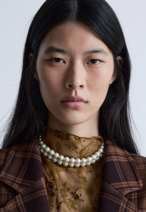 Brown plaid blazer, sheer gold floral blouse, and a multi-strand pearl necklace. Smooth textures, high contrast between layered fabrics.