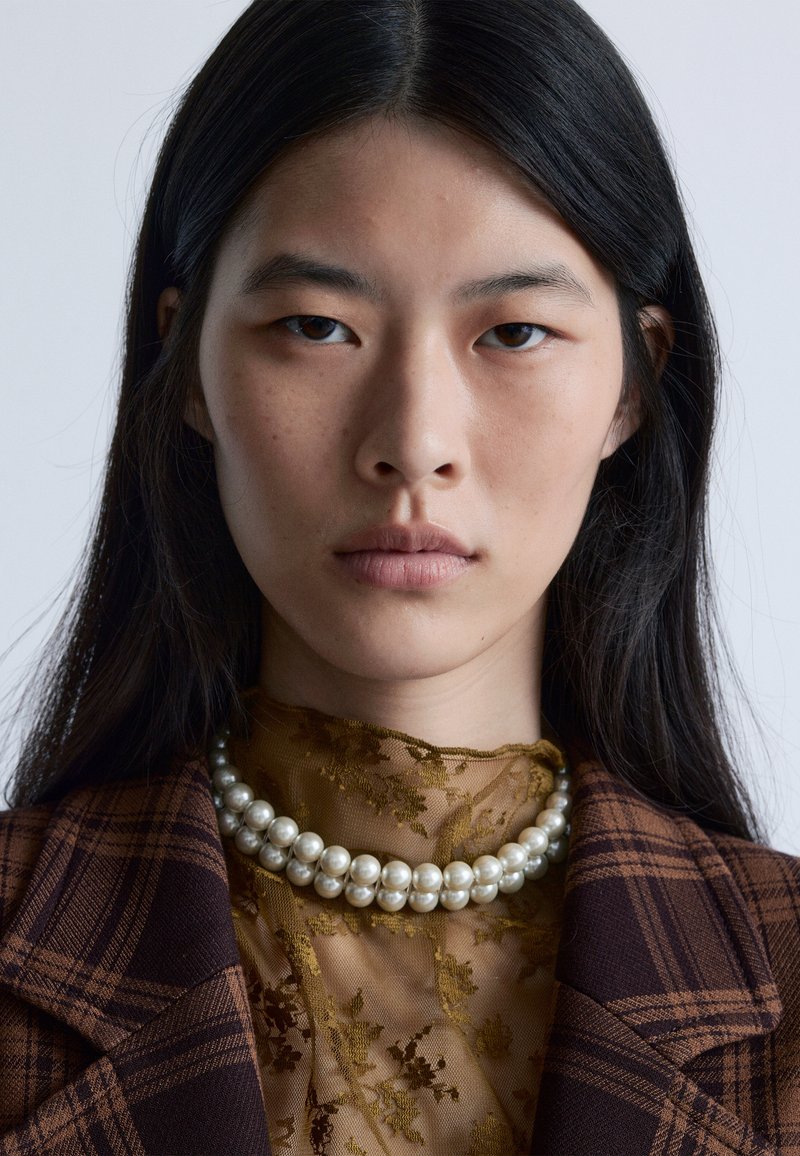 Brauner Karo-Blazer, durchsichtige goldene Bluse mit Blumenmuster und eine mehrsträngige Perlenkette. Glatte Texturen, hoher Kontrast zwischen den übereinander liegenden Stoffen.