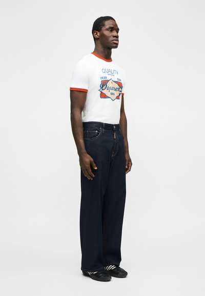 Άντρας που φοράει σκούρο  τζιν με ευρεία ρίγα, λευκό μπλουζάκι με κόκκινη μπορντούρα και μαύρα αθλητικά παπούτσια με λευκές ρίγες, σε απλό γκρι φόντο.