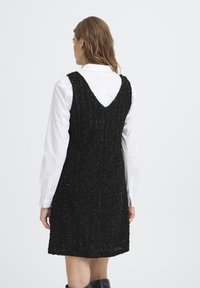 Crna teksturirana haljina s V-oblikom na leđima, koja ima prilagođeni kroj i složen uzorak, nosi se preko bijele košulje s dugim rukavima.