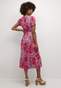 Robe paisley rose avec de courtes manches bouffantes, jupette évasée et taille ajustée. Associée à des sandales à talons épais. Texture lisse.
