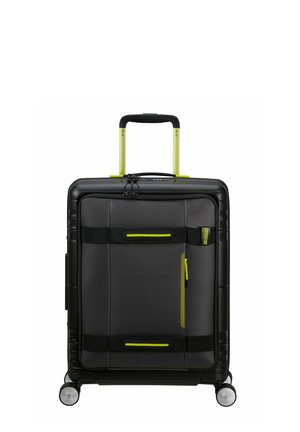 Valise rigide noire avec des accents de fermeture éclair et des poignées vertes, présentant une surface texturée et quatre roues pour la mobilité.