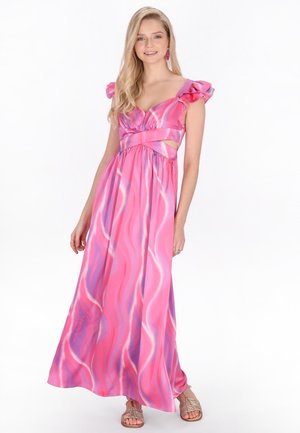 Kobieta ubrana w różowo-fioletową suknię maxi bez rękawów z falistym wzorem, ozdobioną falbankami na ramionach i wycięciami po bokach, do tego złote sandały.