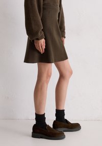 Persoon draagt een bruine wollen jurk, zwarte sokken en donkerbruine loafers, staand tegen een witte, getextureerde muur.